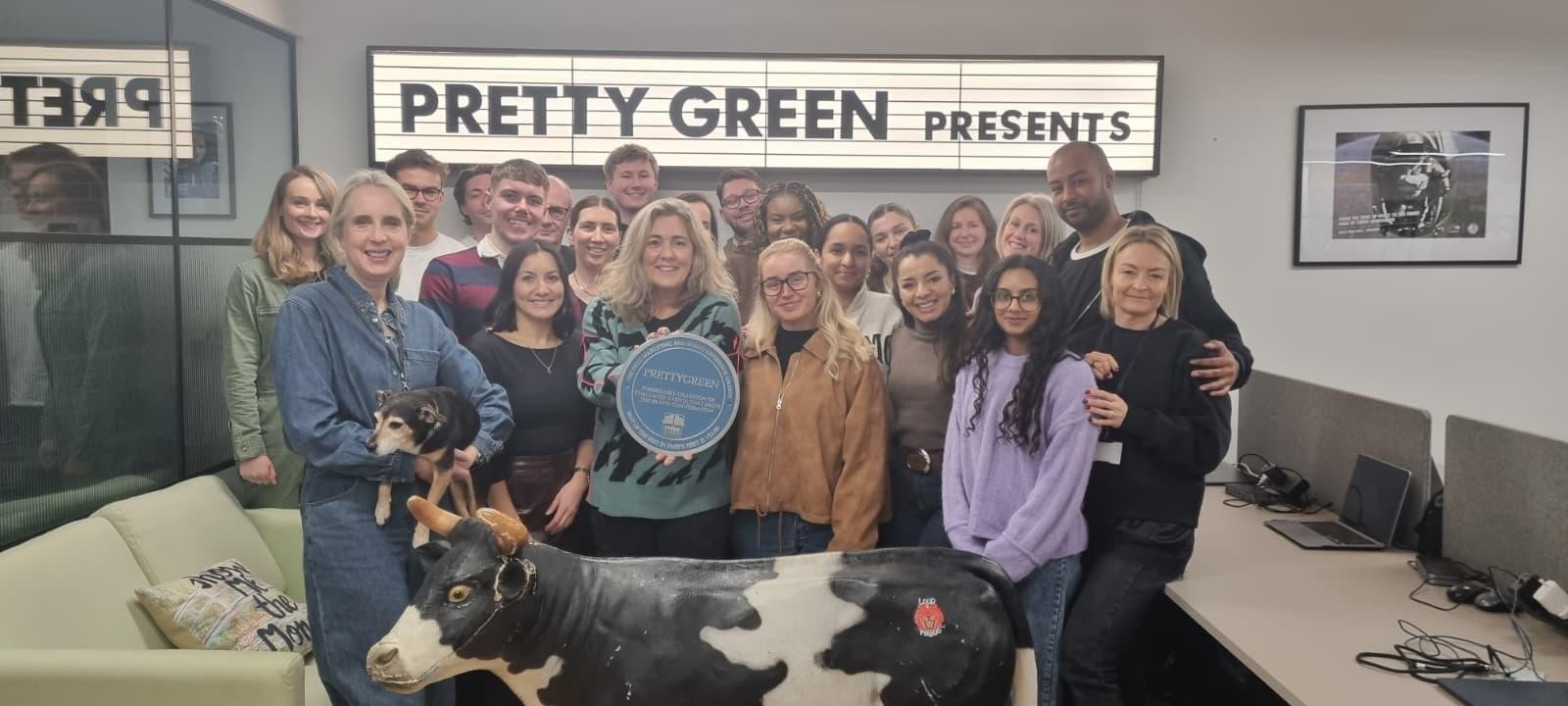 A diverse group of smiling individuals poses together in a modern office, with a sleek glass wall behind them. In the foreground, a black-and-white cow figurine adds a quirky touch, while a neon sign reads 'PRETTY GREEN PRESENTS'. Bright office lighting enhances the welcoming atmosphere.