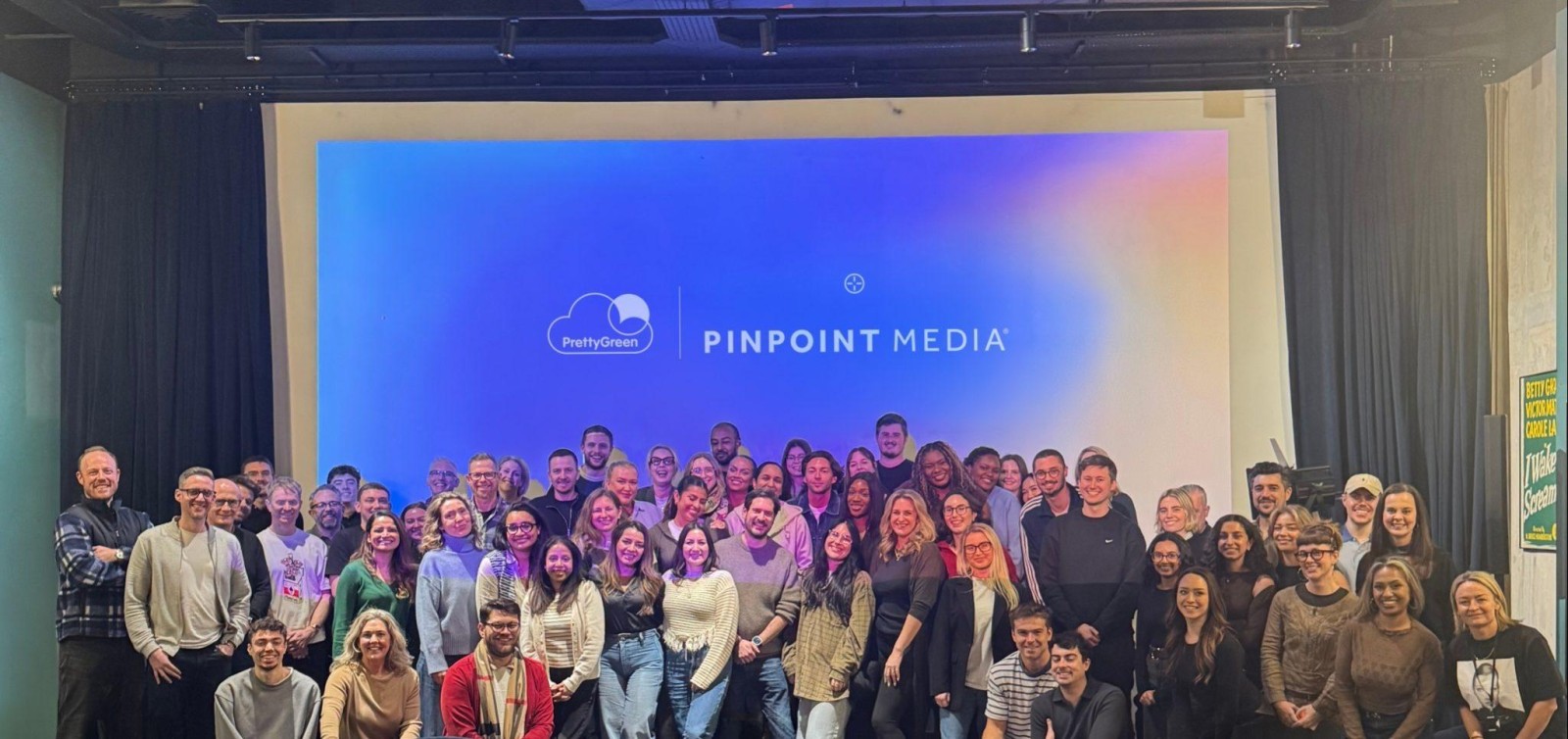 A large group photo featuring the diverse team at Pinpoint Media, standing in front of a vibrant blue and purple backdrop with the company logo. The atmosphere is lively and energetic, as individuals display a range of casual attire, contributing to a sense of camaraderie.