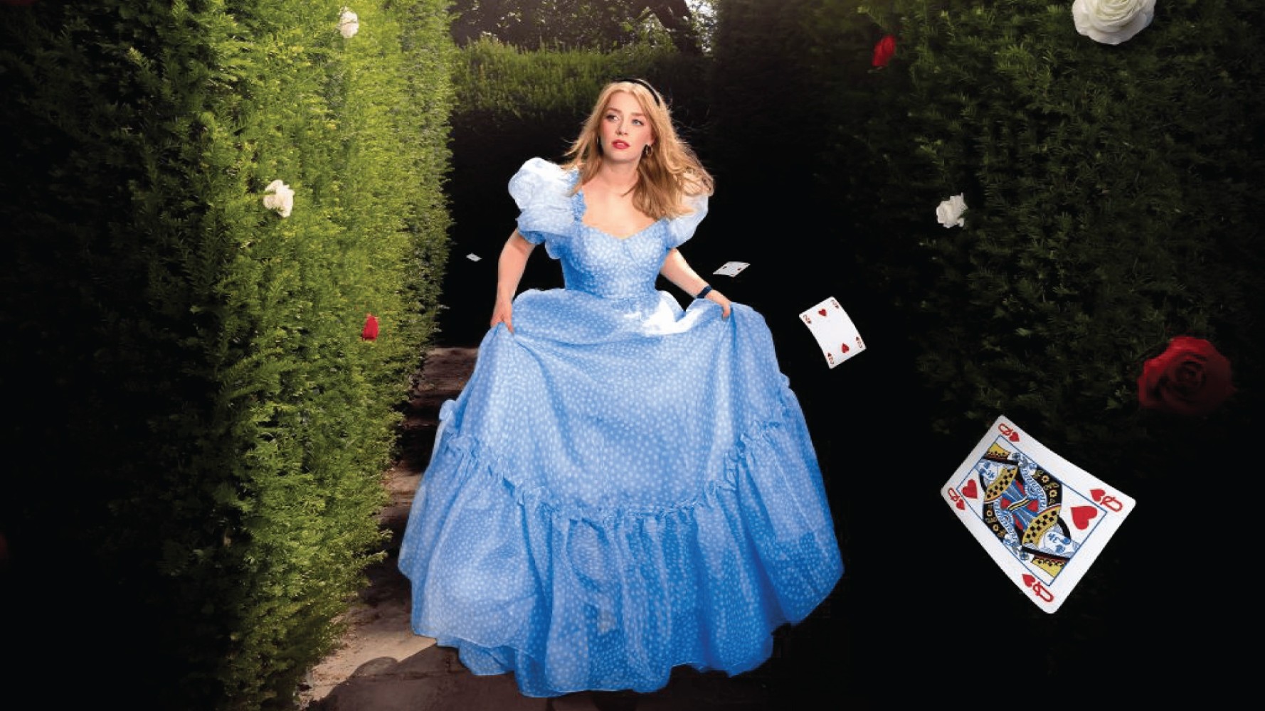 A woman in a flowing sky-blue gown, adorned with delicate white polka dots, runs along a narrow stone path flanked by lush, green hedges. Playing cards, notably a queen and a king, flutter down elegantly around her, while blooming roses add vibrant pops of red and white to the enchanting scene.