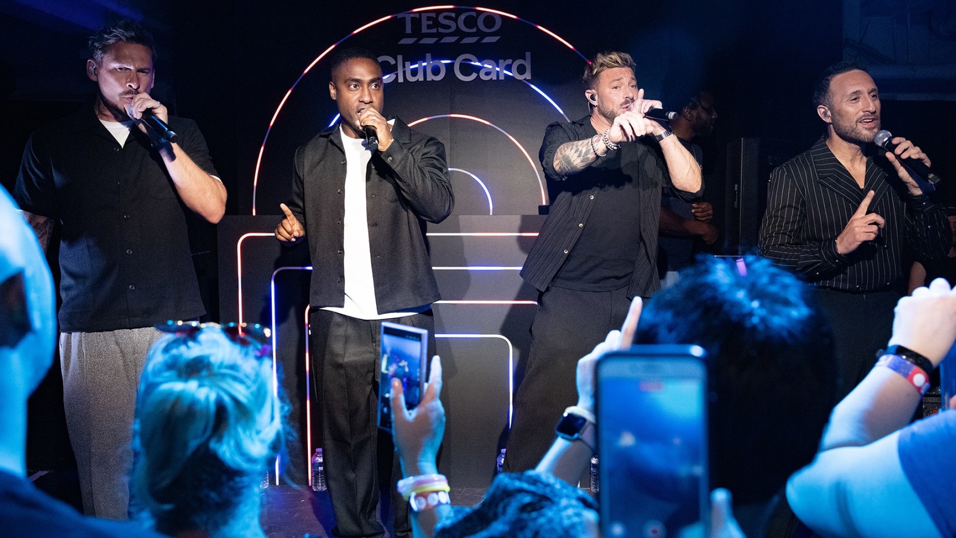 Four male singers stand on stage, captivated in a performance. The lead vocalist, dressed in a black shirt, holds a microphone, while others, in stylish outfits, harmonise passionately. Behind them, vibrant lights illuminate the venue, creating an energetic atmosphere filled with anticipation.