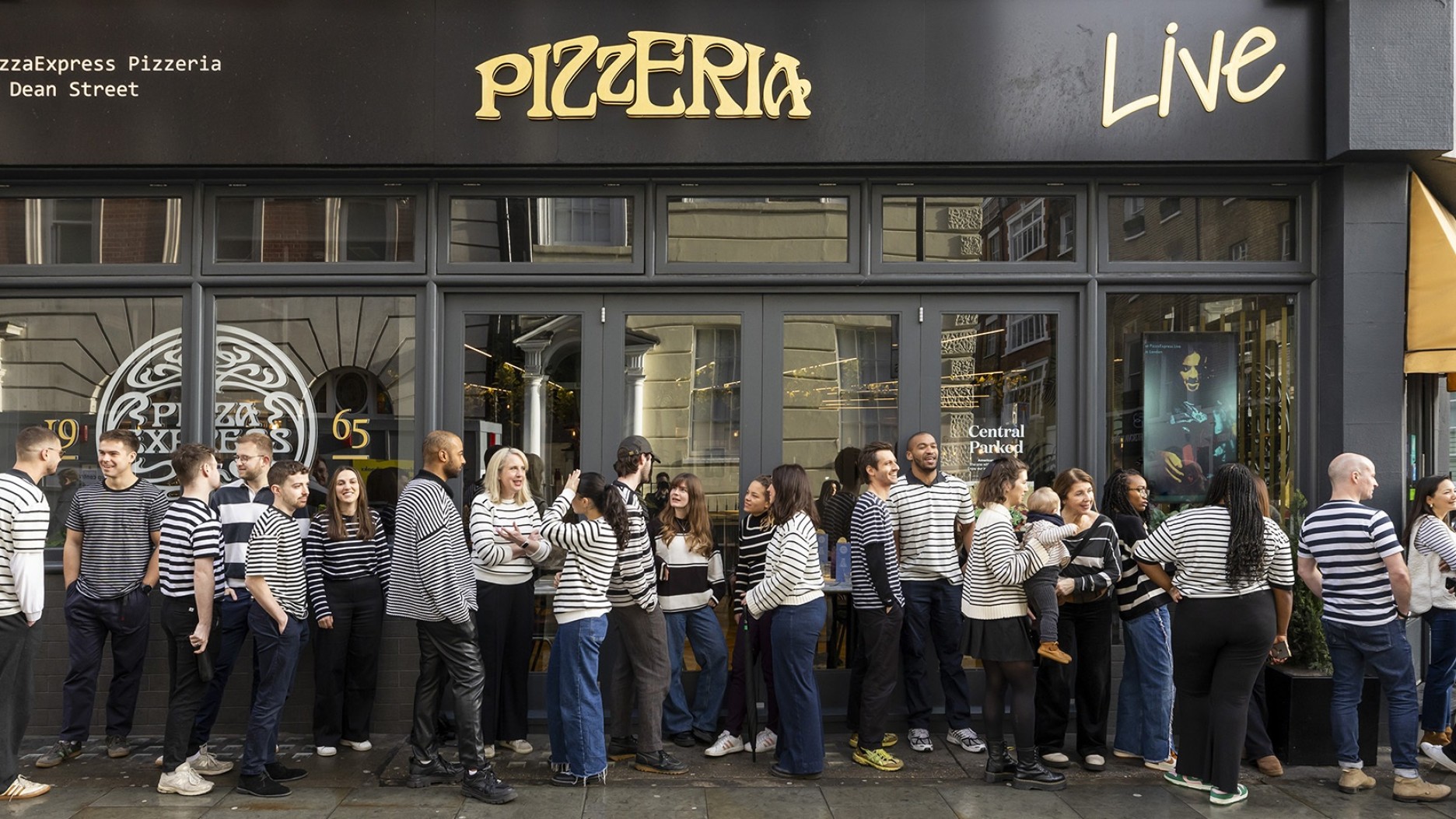 A diverse group of people wearing striped shirts stands outside Pizzexpress Pizzeria, showcasing a playful spirit. The sleek, modern shop front features large glass windows and an inviting canopy, while natural light enhances the vibrant atmosphere of the bustling street.