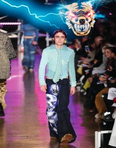 A young male model strides confidently down the runway, showcasing an eye-catching outfit. He wears a light blue, textured jacket layered over a black ensemble embellished with intricate tiger designs. A playful, graphic backdrop of a roaring tiger's face, surrounded by electric blue light, adds an edgy flair to the vibrant atmosphere of the fashion show.