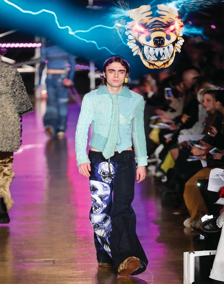 A young male model strides confidently down the runway, showcasing an eye-catching outfit. He wears a light blue, textured jacket layered over a black ensemble embellished with intricate tiger designs. A playful, graphic backdrop of a roaring tiger's face, surrounded by electric blue light, adds an edgy flair to the vibrant atmosphere of the fashion show.