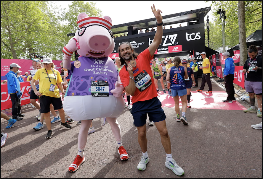 A vibrant scene at a marathon finish line, featuring a cheerful man in an orange shirt and navy shorts posing alongside a large pink pig mascot adorned with a purple vest promoting the National Deaf Children’s Society. In the background, a lively crowd of runners and supporters gather under bright blue skies, creating an atmosphere of celebration and camaraderie.