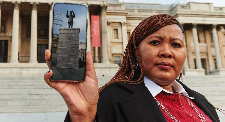 A woman in a black coat holds her smartphone at arm's length, displaying a digital image of a statue on a stone pedestal. She stands on the steps of a grand colonial building with tall columns, while soft sunlight highlights her features, lending a serene quality to the scene.