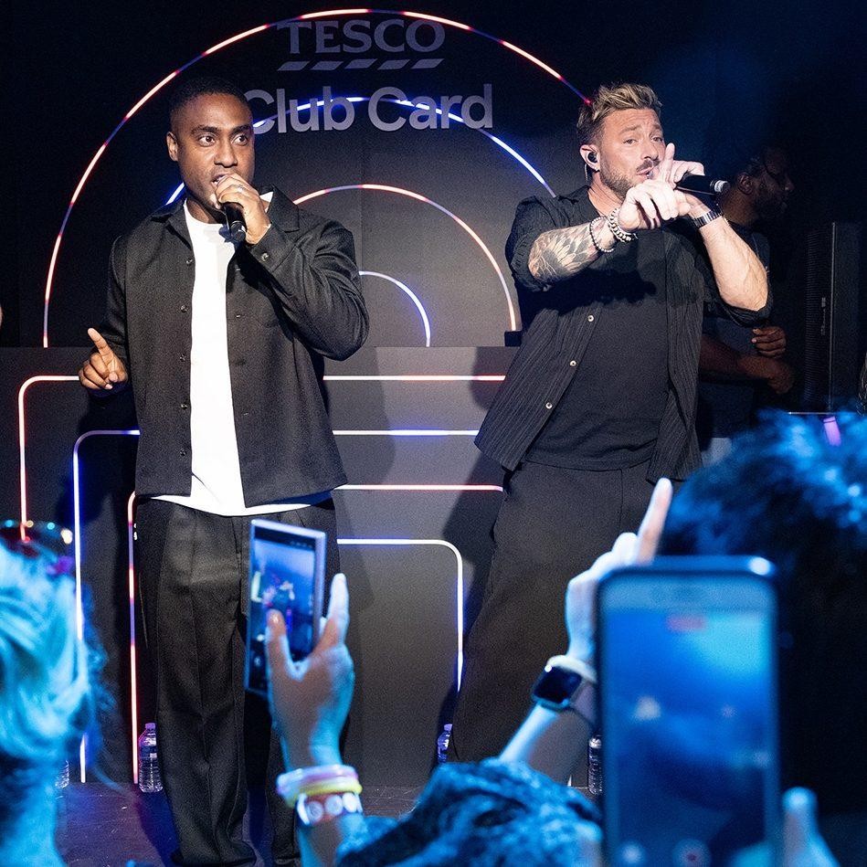 Two male performers take centre stage in a vibrant setting, illuminated by dynamic lighting. One wears a stylish black outfit, microphone in hand, while the other sports a casual black shirt. In the background, neon outlines spell 'Tesco Club Card', enhancing the energetic atmosphere of the live performance.