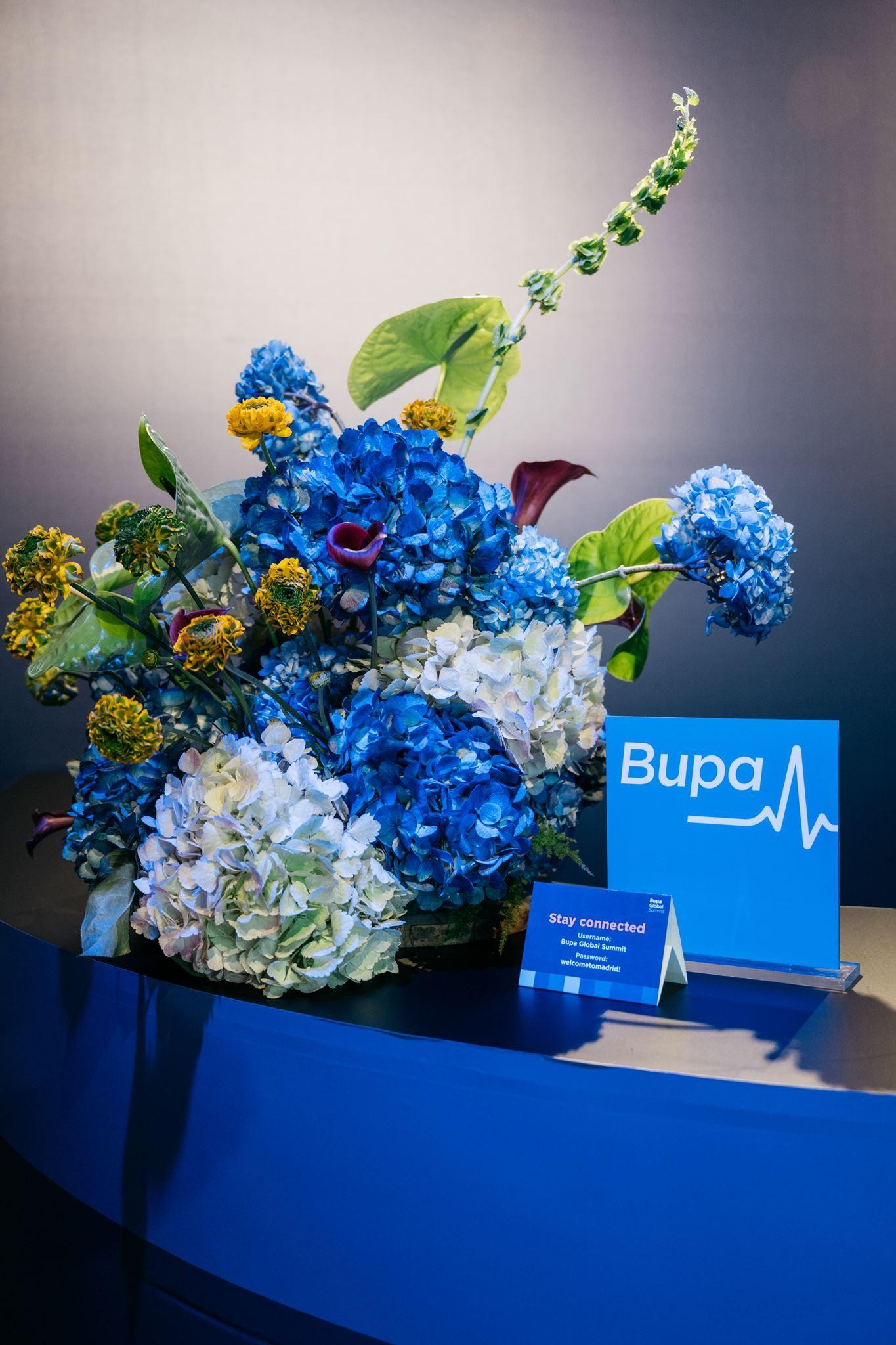 An elegant display of flowers features vibrant blue hydrangeas, accented by yellow florals and lush green leaves, forming a striking focal point. In front, a bright blue sign with the Bupa logo and an invitation to stay connected rests on a sleek counter, enhancing the modern aesthetic.