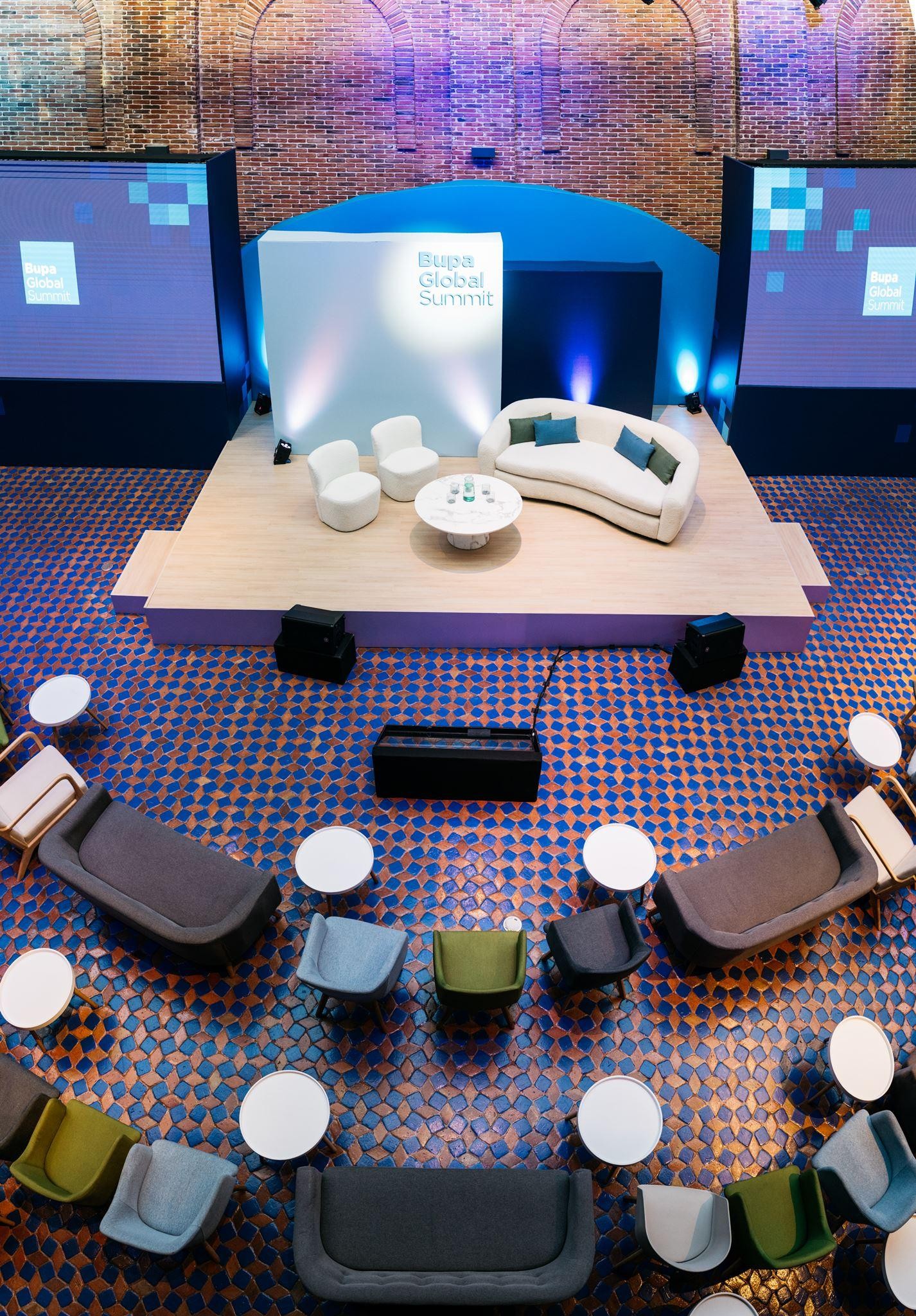 An overhead view of an elegantly arranged conference space for the Blage Global Summit. A sleek, curved white sofa forms a focal point on a pale wooden stage, surrounded by low white tables. Comfortable seating in varying shades of grey and green is positioned in a semi-circle, enhancing the inviting atmosphere. Soft blue and white lights illuminate the brick walls, creating a modern yet warm environment.