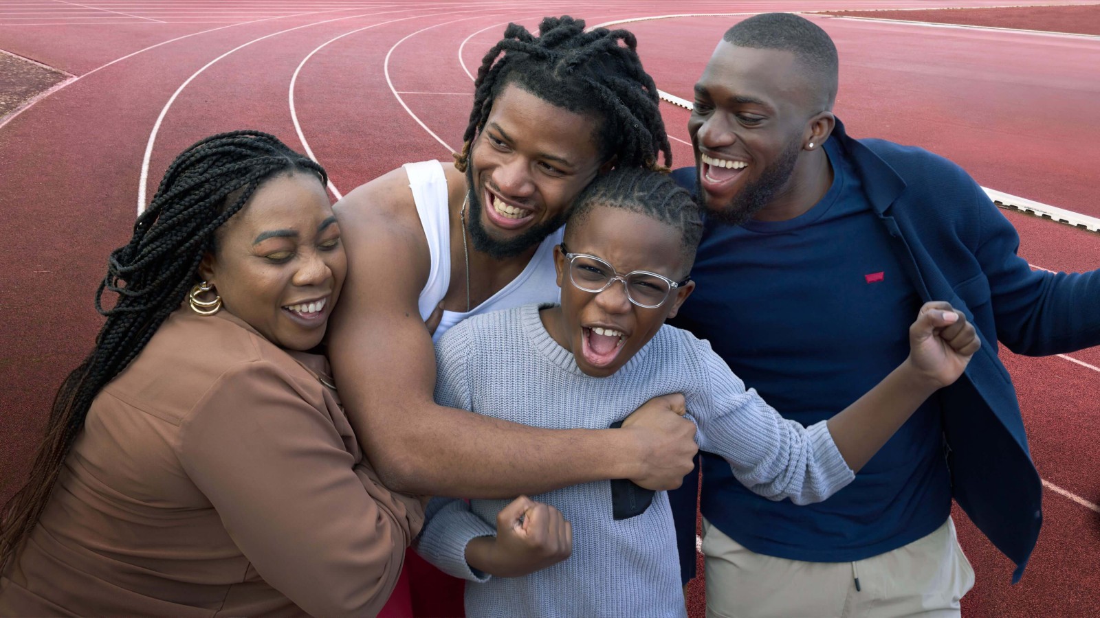 Four people gather joyfully on a vibrant red athletic track, embracing and laughing. A woman in a brown blouse stands to the left, her hair in waves, while a man in a white tank top is beside her, showcasing his strong build. A young boy in a grey sweater beams enthusiastically, hugged tightly by another man in a navy jacket. The sun casts a bright light, enhancing the lively atmosphere.