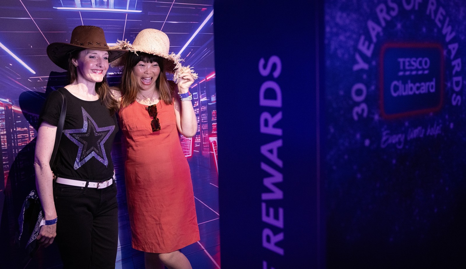 Two women pose playfully in a vibrant setting adorned with neon lights, wearing oversized hats. One wears a black shirt with a star graphic and jeans, while the other dons a bright orange dress. A bold display celebrating 30 years of Tesco Clubcard is visible, enhancing the festive atmosphere.