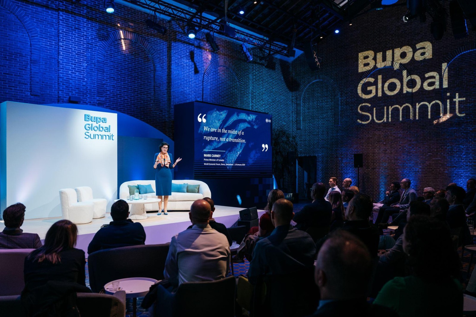 An engaging speaker stands on stage at the Bupa Global Summit, illuminated by soft spotlights. Behind her, a large screen displays quotes, framed by an elegant blue backdrop. Attendees sit in a semi-circle on plush sofas, attentively listening in a stylish, contemporary venue with exposed brick walls.