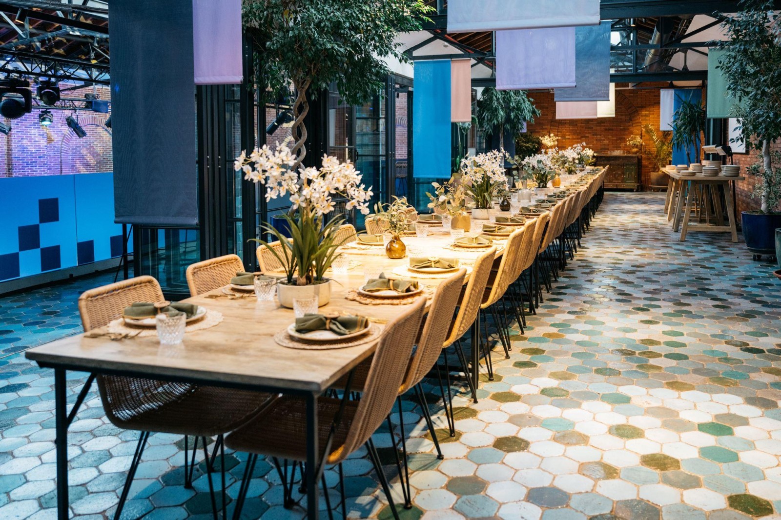 A long wooden dining table elegantly set in a spacious, modern venue, adorned with delicate white orchids and glass tableware. The floor features colourful hexagonal tiles, while soft light filters through a canopy of greenery, creating a serene, inviting atmosphere.
