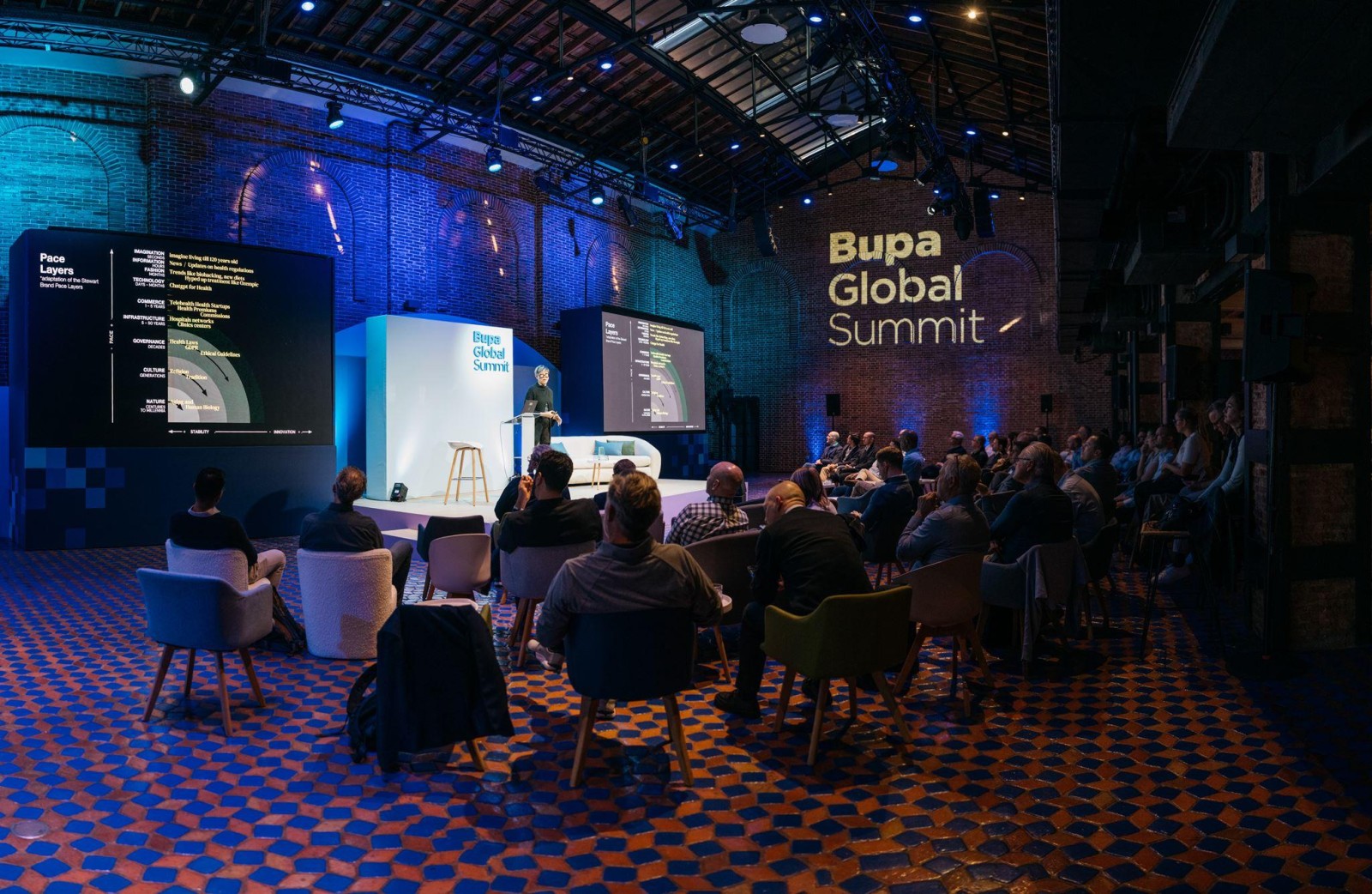A spacious venue with an industrial atmosphere features a presentation stage adorned with a large screen displaying digital content. Attendees, seated in modern white and colourful armchairs, absorb insights during the Bupa Global Summit. Soft blue lighting accentuates the textured brick walls, creating a sophisticated ambiance.