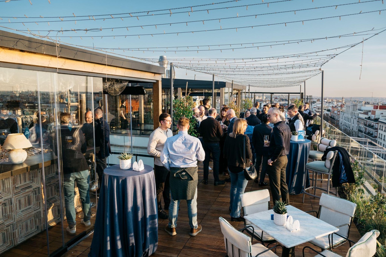 A lively rooftop gathering with a panoramic city skyline in the background. Guests are mingling around elegant round tables draped in navy cloth, accompanied by modern chairs. String lights twinkle above, creating a warm atmosphere in the evening light.
