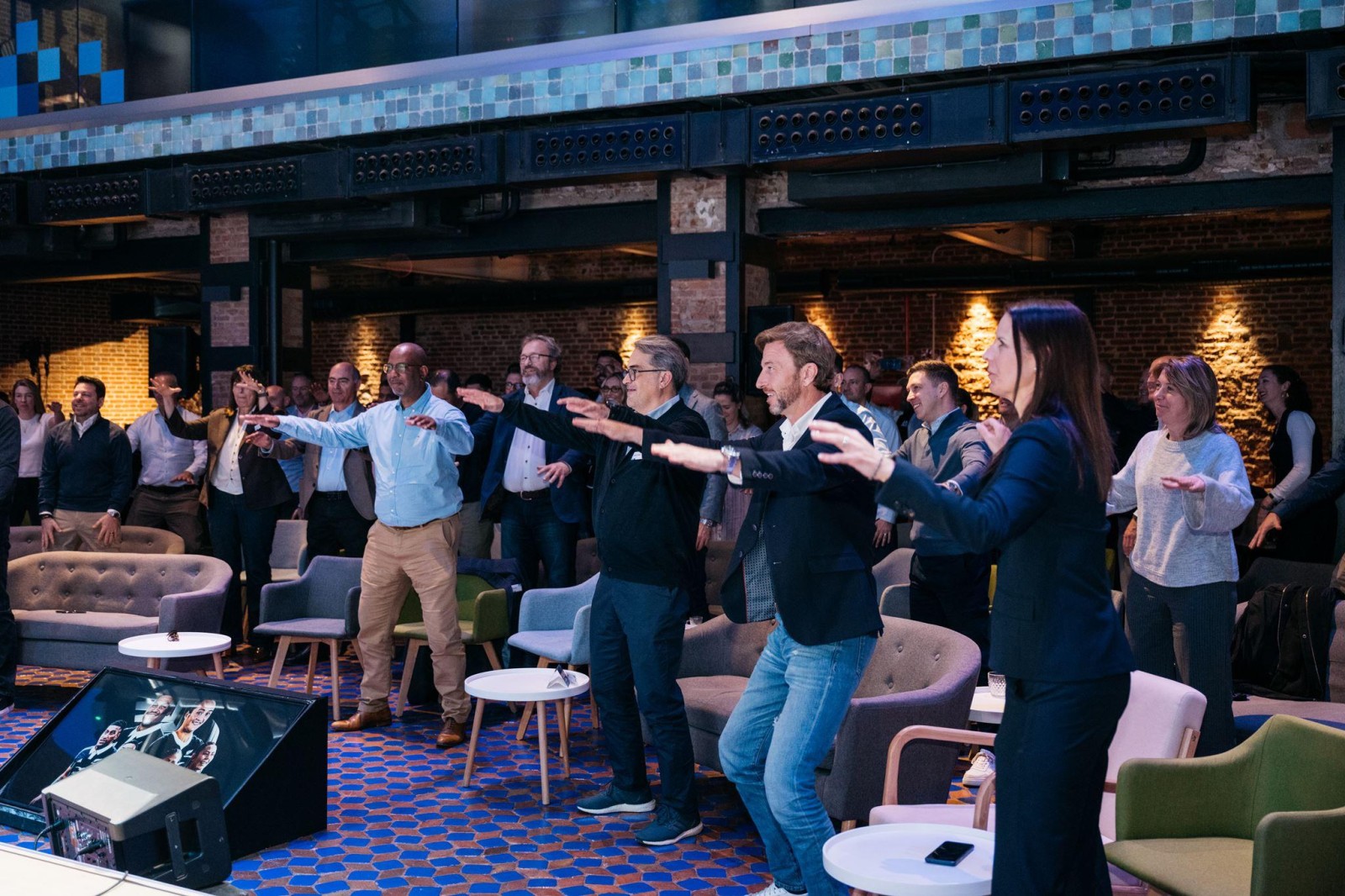 A diverse group of adults, both men and women, stand with arms extended in a lively, interactive setting, surrounded by modern furnishings. They are engaged in a dynamic activity, with a backdrop of brick walls and ambient lighting casting a warm glow across the room.
