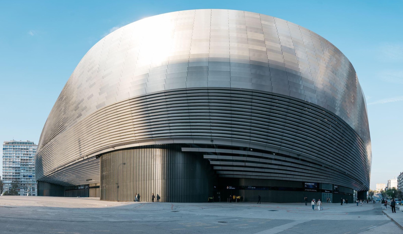 A striking modern arena features a polished metallic exterior with smooth, curved lines and horizontal grooves, creating a dynamic visual flow. Large glass panels reflect sunlight, while pedestrians appear small against the expansive plaza, highlighting the building's grandeur.