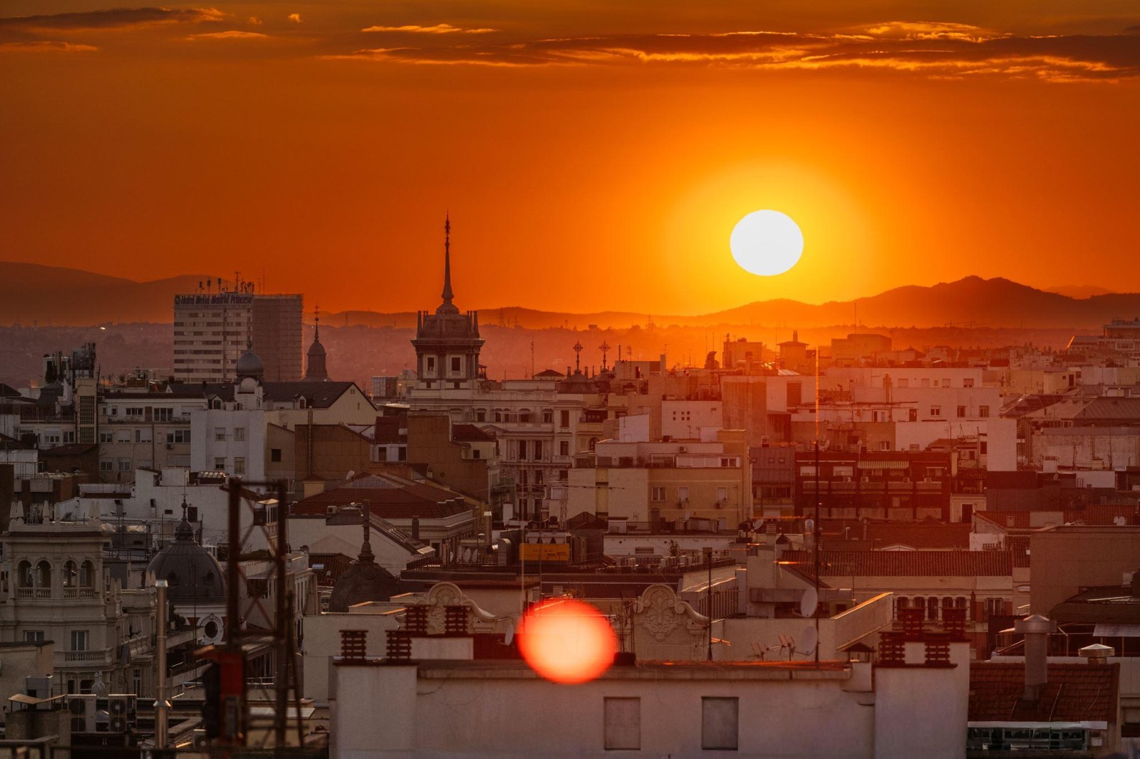 A breathtaking sunset over a sprawling cityscape, with the sun's golden orb sinking behind distant mountains. Buildings, bathed in warm orange hues, create a textured skyline, interspersed with steeples and rooftops. The soft glow contrasts against the fading daylight, evoking a serene atmosphere.