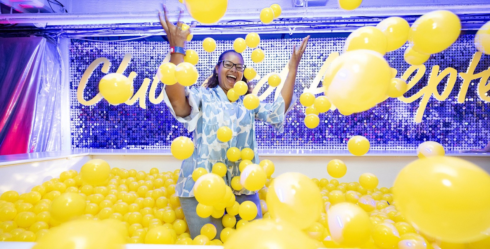 A joyful woman dressed in a light blue blouse stands amidst a vibrant sea of yellow balloons in a bright, playful setting. The background features sparkling sequins that shimmer under soft lighting, enhancing the festive atmosphere. The space is filled with a sense of exuberance and celebration.