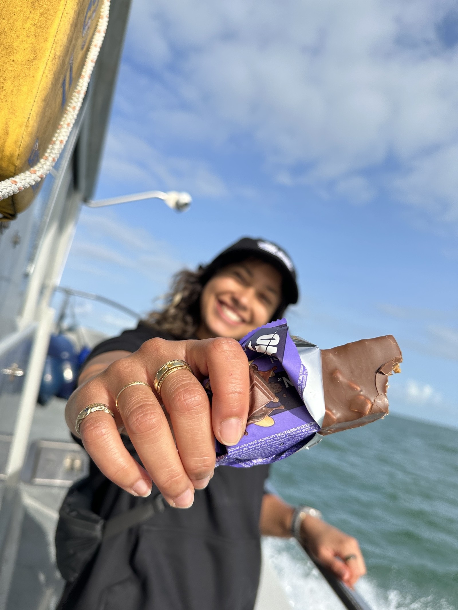 A woman with curly hair, wearing a black cap and a loose black top, stands on a boat's deck, proudly offering a partially eaten chocolate bar wrapped in purple packaging. The shimmering sea glistens beneath a bright blue sky dotted with fluffy clouds, creating a vibrant, joyful atmosphere.