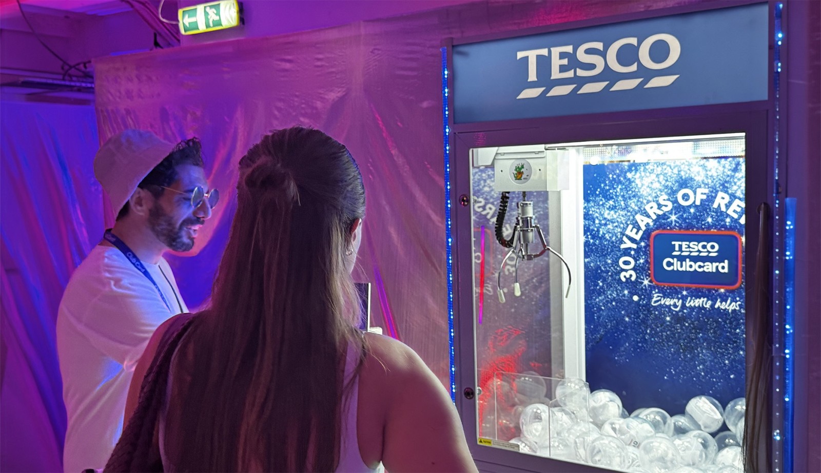 A man wearing sunglasses and a white cap stands beside a woman with long hair, both focused on a vibrant claw machine emblazoned with the Tesco logo. The machine is illuminated with blue LED lights, surrounded by a soft, purple backdrop, creating a festive atmosphere.