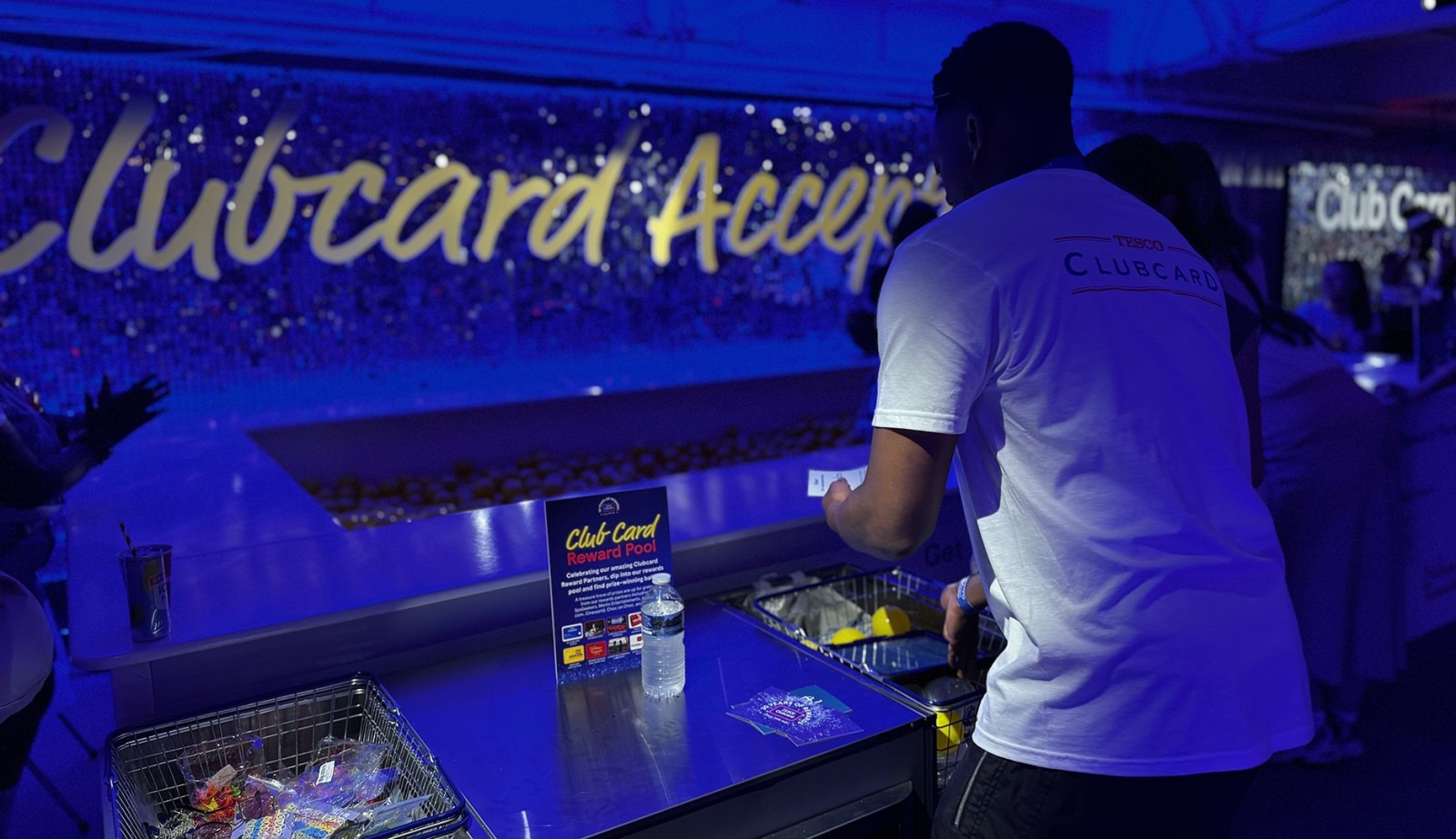 A vibrant scene featuring a young man in a white t-shirt at a sleek service counter, illuminated by soft blue lights. The background shows shimmering decorations, enhancing the festive atmosphere. The counter is adorned with a clear bottle of water and promotional materials for a loyalty programme, while a group of hands reach towards enticing treats.