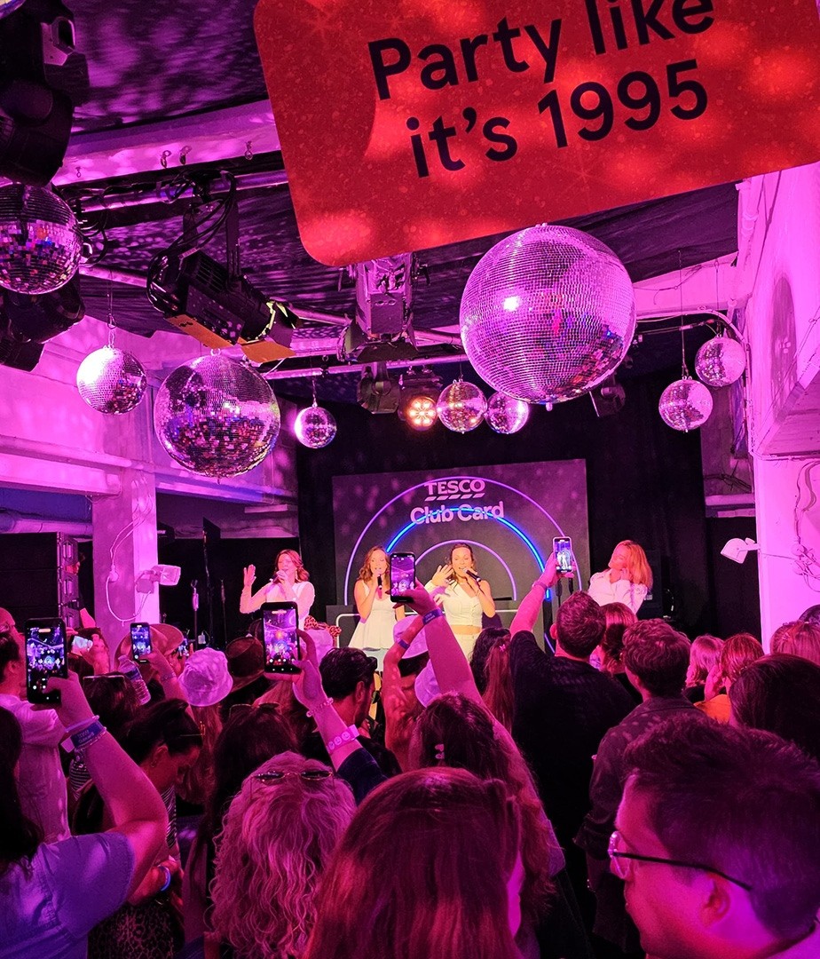 A lively event scene filled with people enjoying a performance under shimmering disco balls. The stage features vibrant pink lights, a backdrop displaying 'TESCO Club Card,' and attendees capturing the moment on their phones, all exuding a nostalgic 1990s party mood.