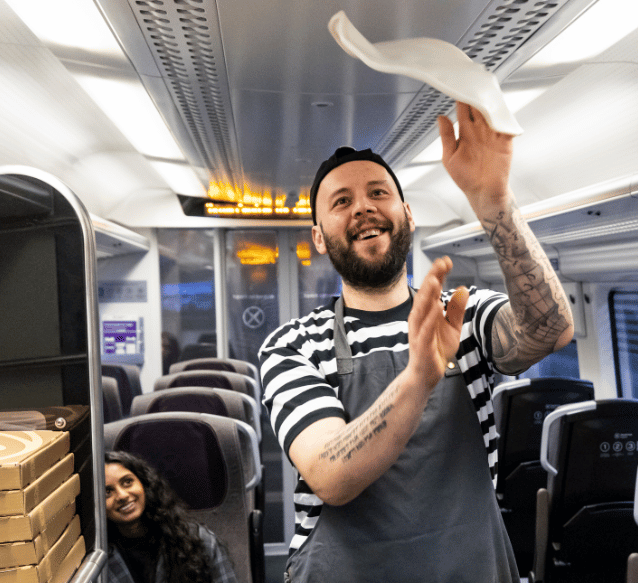 A chef for PizzaExpress is on a train in a stripey tshirt and apron tossing a pizza with his hands