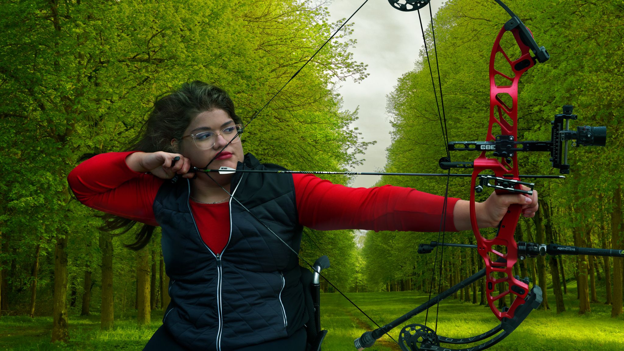 A woman with glasses, Paralympian Mariana Zúñiga is outdoors in a green forest, focusing intently on a target while holding a red compound bow in full draw position. She is wearing a black sleeveless jacket over a long-sleeved red shirt.