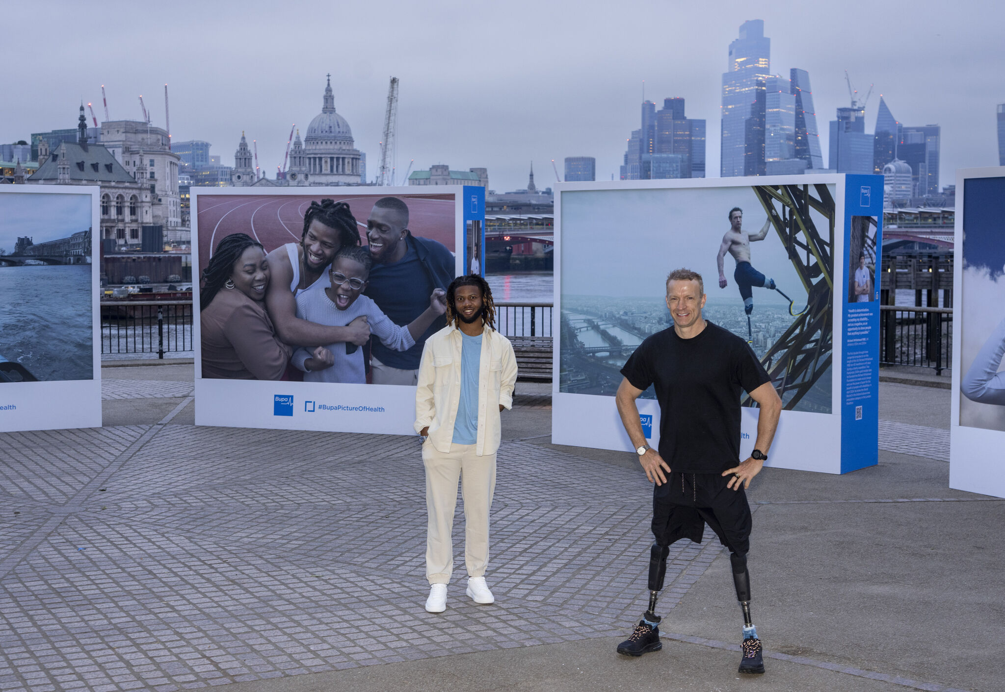 Two men stand on London's Southbank infront of an open air gallery, one of the men has prosthetic legs.