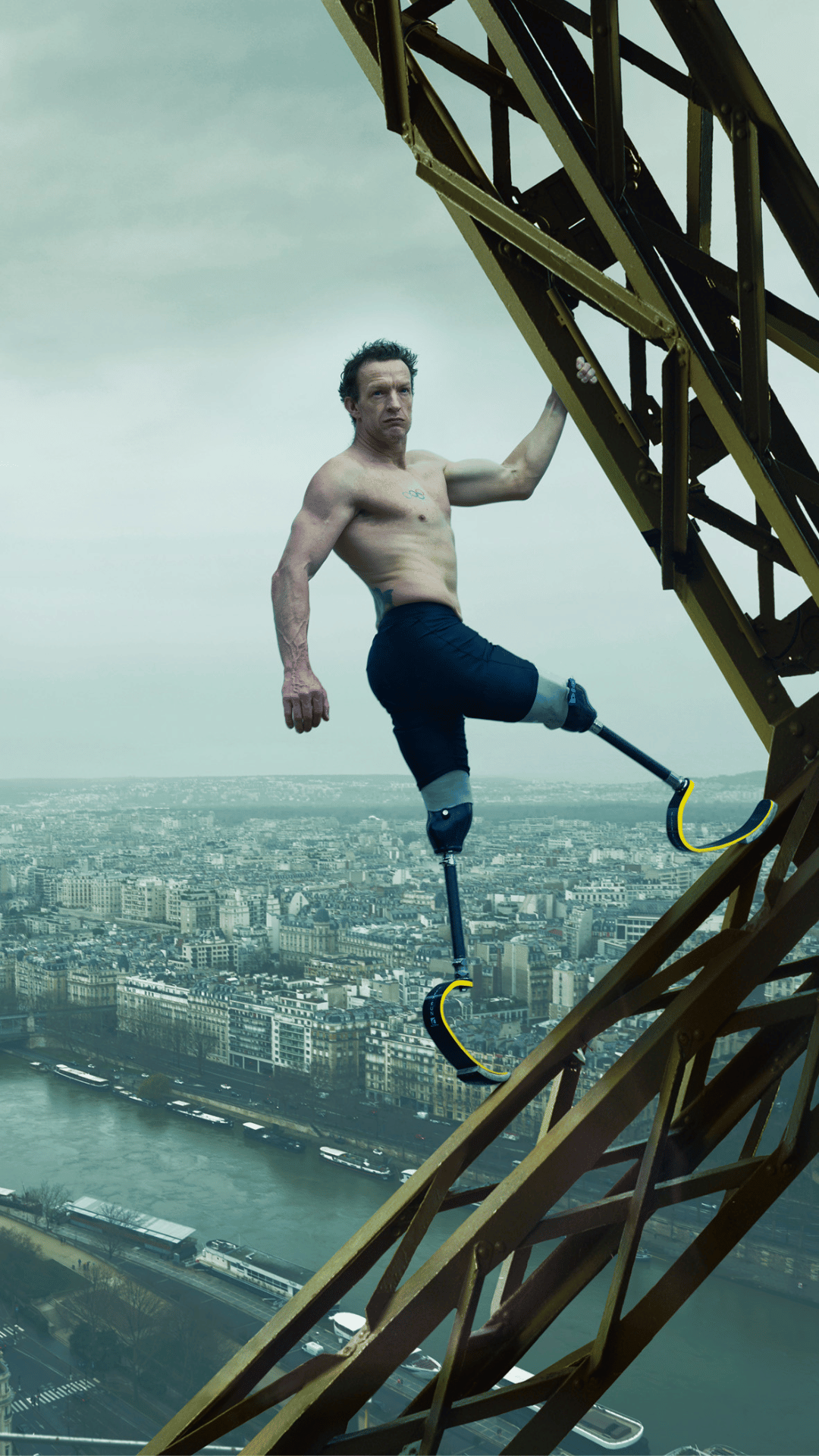 An image of UK Paralympian Richard Whitehead MBE, shirtless with prosthetic running blades is climbing a tall metal structure, with an urban cityscape visible in the background.