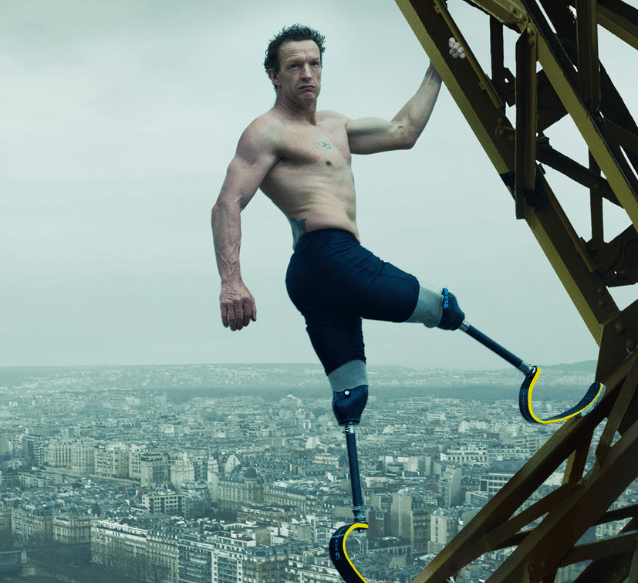 An image of UK Paralympian Richard Whitehead MBE, shirtless with prosthetic running blades is climbing a tall metal structure, with an urban cityscape visible behind him.