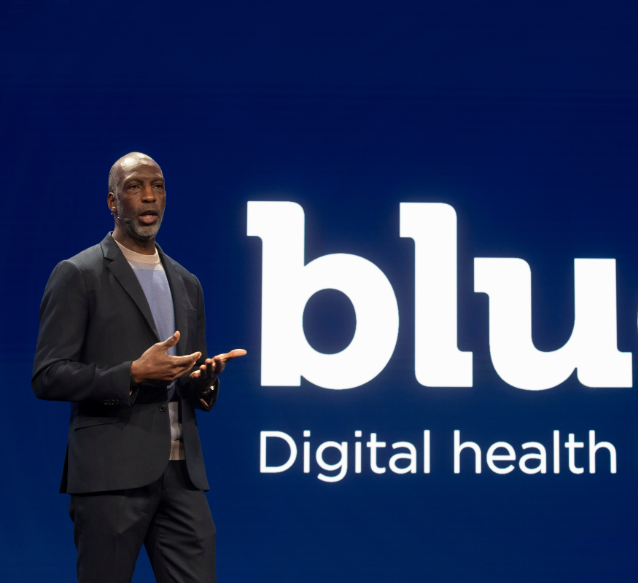 A person in a suit, Michael Johnson, is speaking on a stage. Behind them, a large screen displays the text