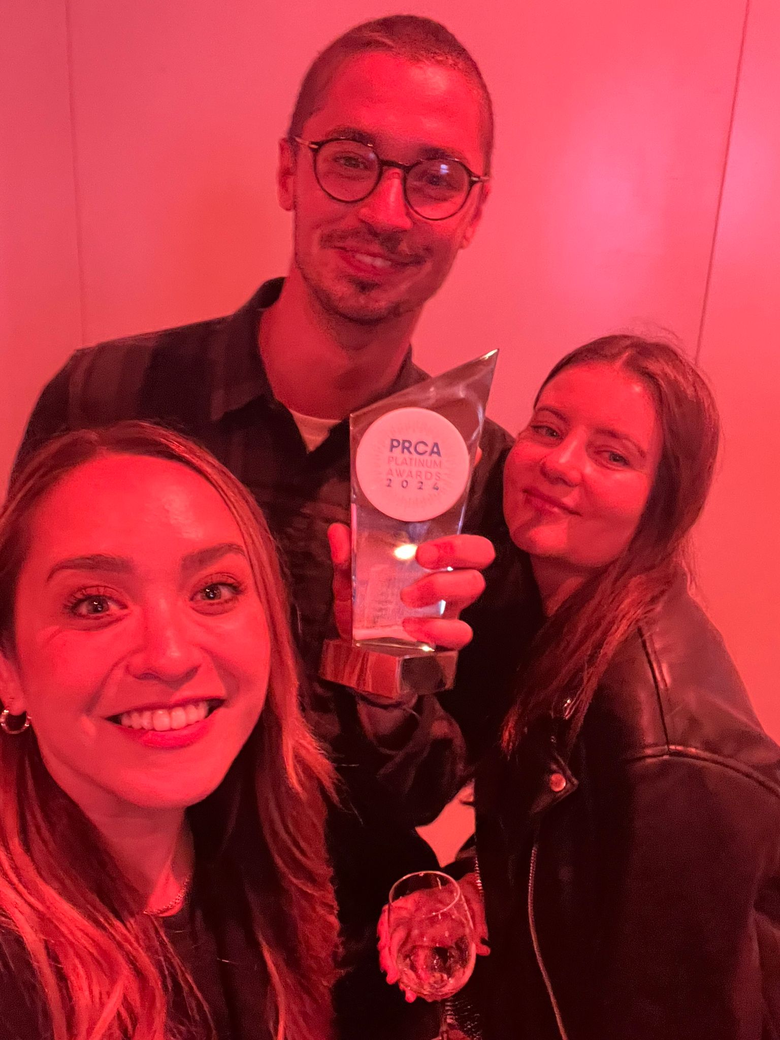 A man is stood smiling wearing glasses holding an award up between two smiling women