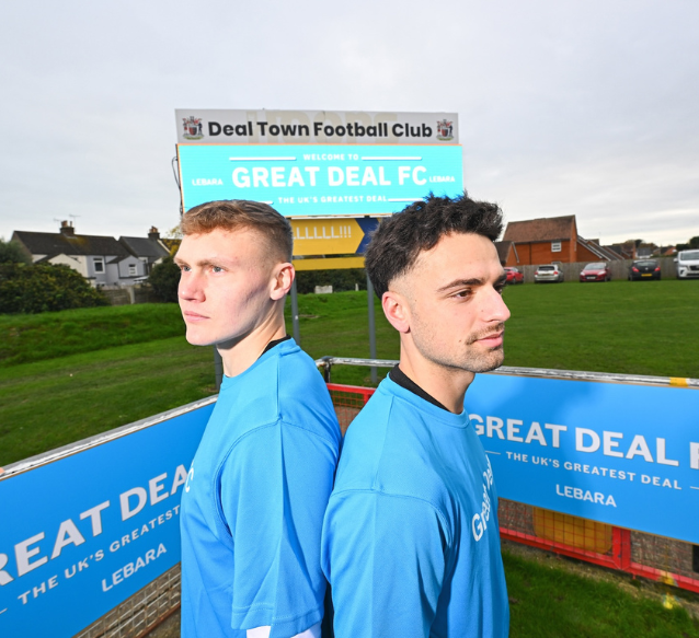 Two men are stood back to back wearing blue football shirts by a pitch