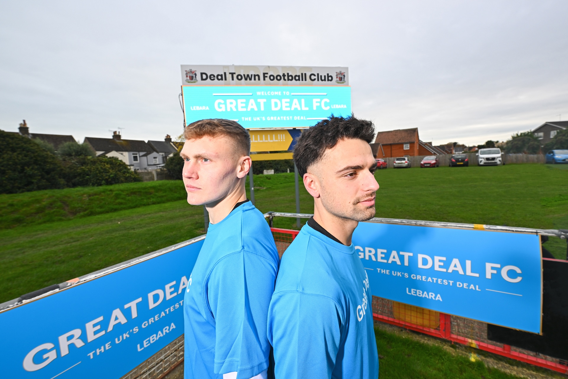 Two men are stood back to back wearing blue football shirts by a pitch