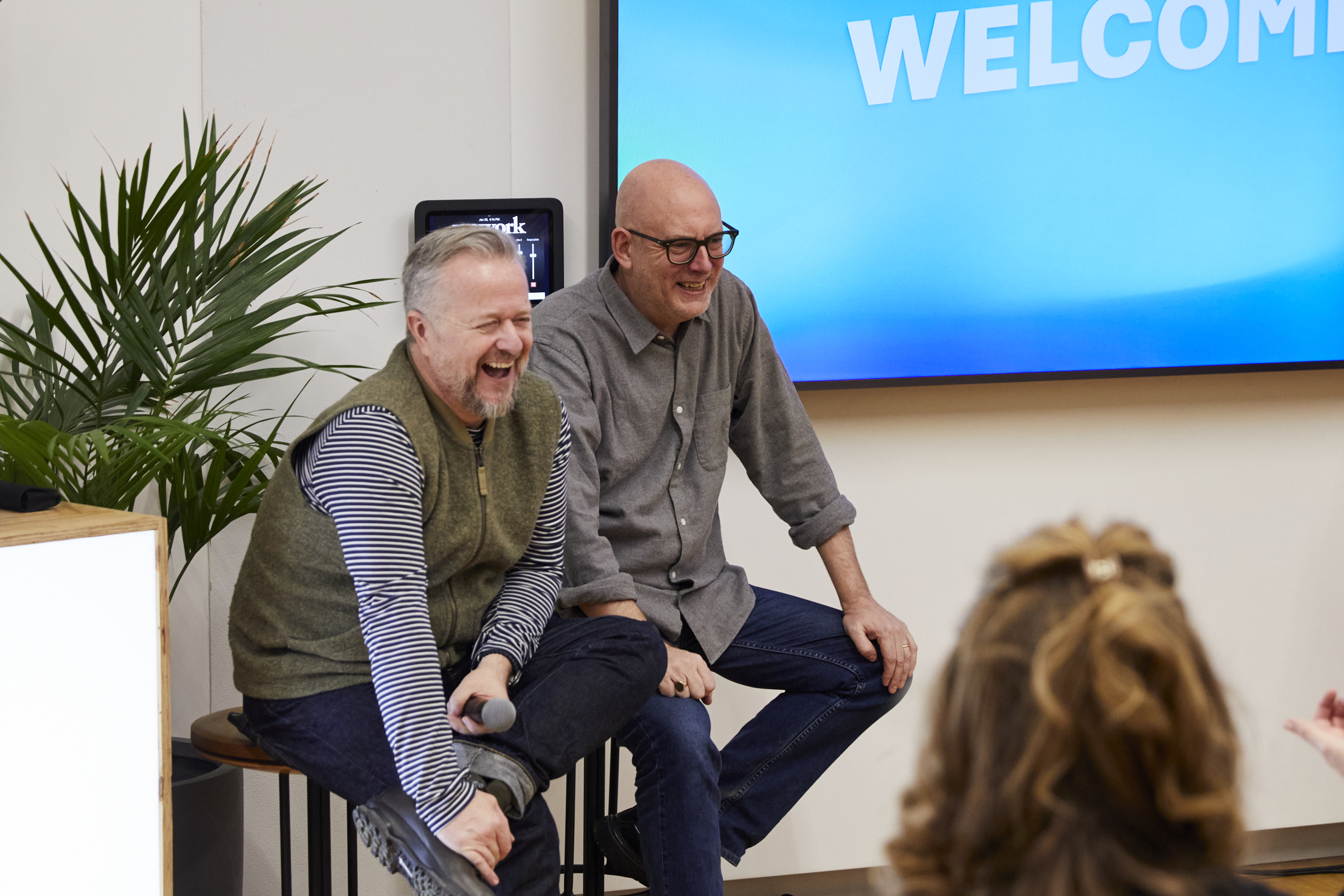 Two men on stools are laughing in front of an audience