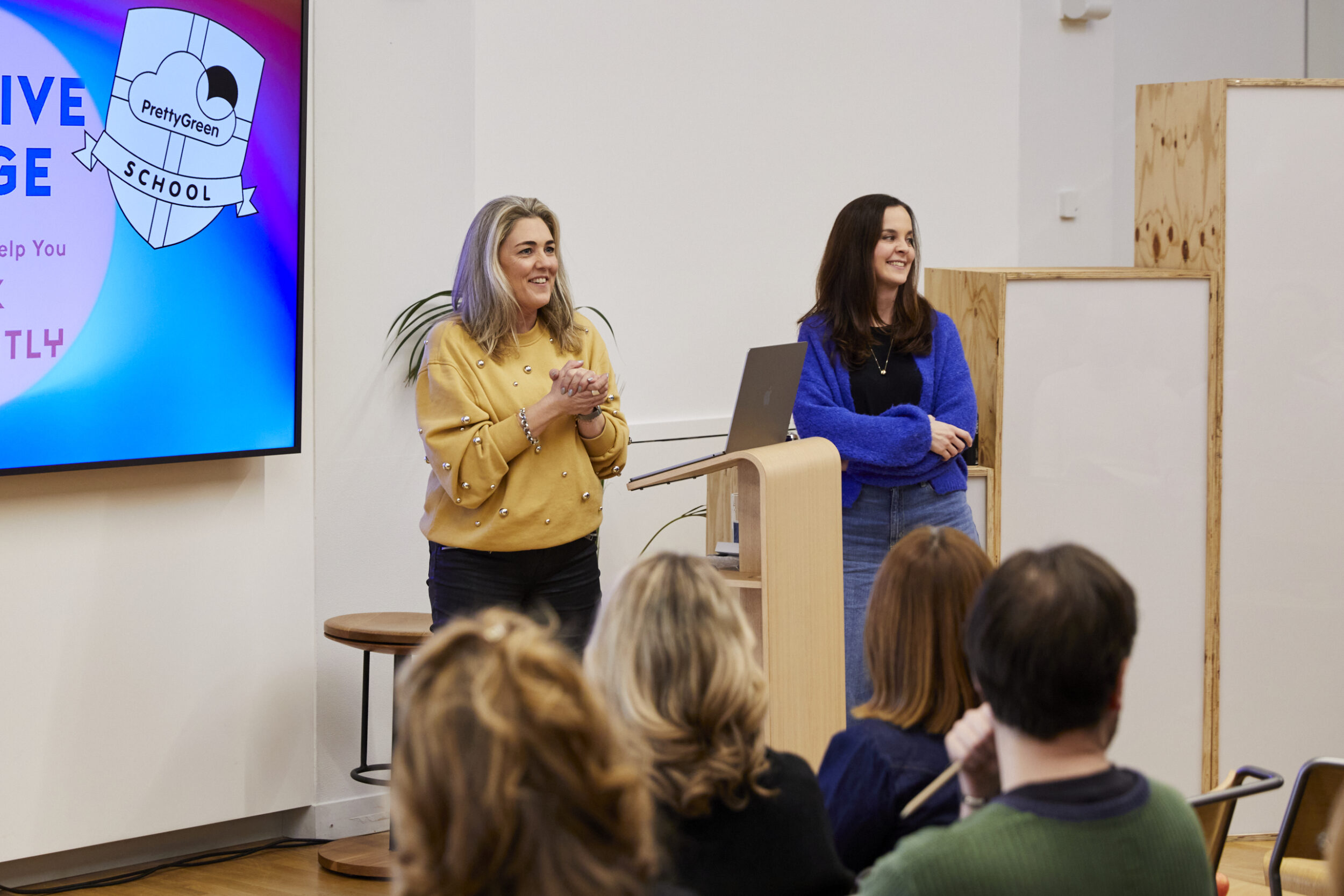 A women in a yellow jumper and one in blue are stood animated in front of a group of people